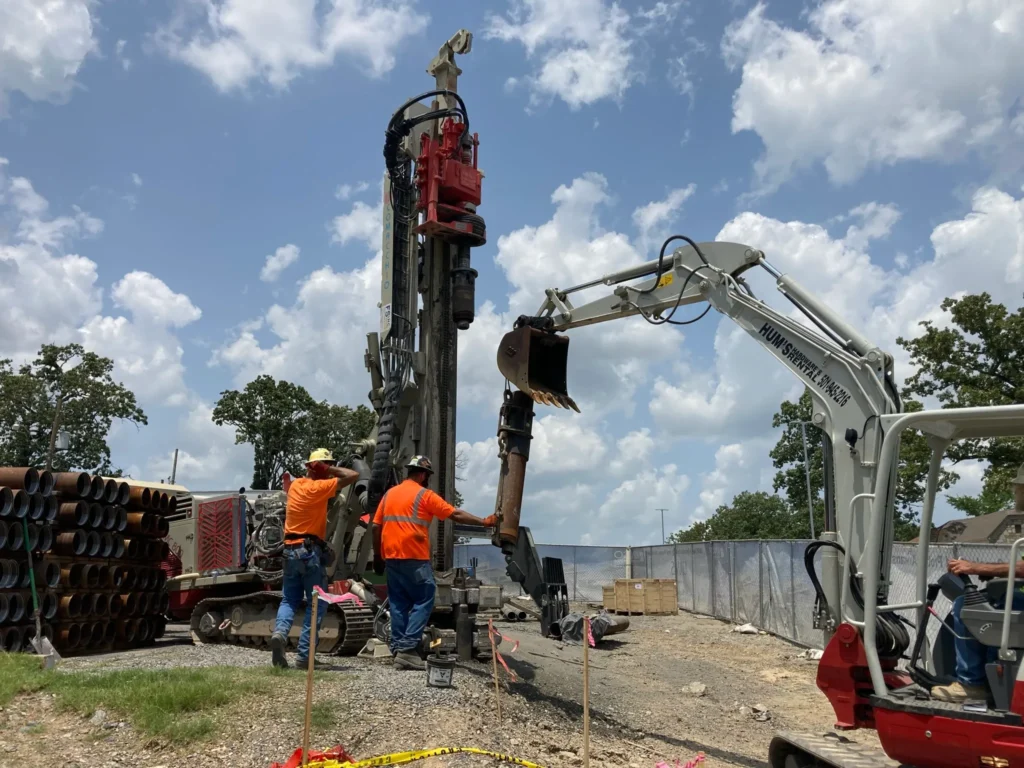 Construction workers operate heavy machinery at a job site. One worker uses a large drilling rig while another operates an excavator. Metal pipes are stacked nearby, and trees are in the background under a partly cloudy sky.