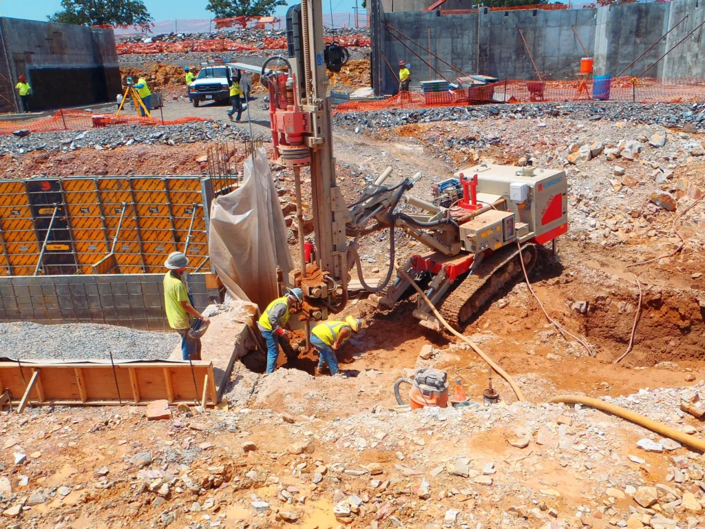 Construction site with workers operating heavy machinery, drilling into the ground. The area is surrounded by safety barriers and mixed terrain, and other workers are visible in the background, near partially constructed structures and equipment.