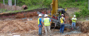 Construction workers in safety gear operate heavy machinery at a wooded construction site. They stand on cleared earth, surrounded by trees and materials, engaged in a project.