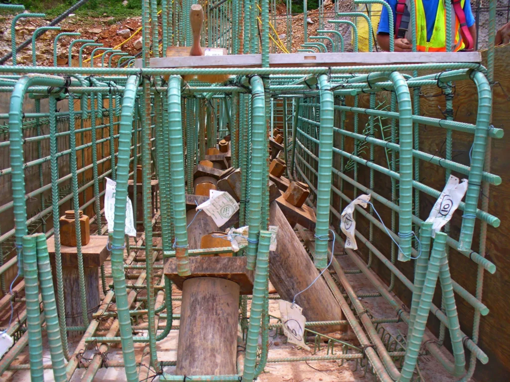 Close-up of a construction site showing green steel rebar arranged in a grid pattern for concrete reinforcement. Wooden formwork and tagged wires are visible, with a worker in a high-visibility vest in the background.