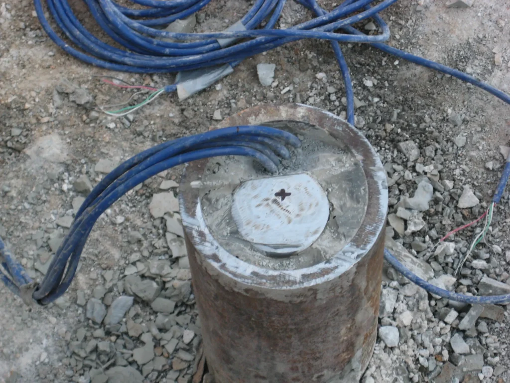 Close-up of a construction site showing a concrete pillar with several blue cables emerging from its top. The ground around the pillar is covered in gravel and debris.