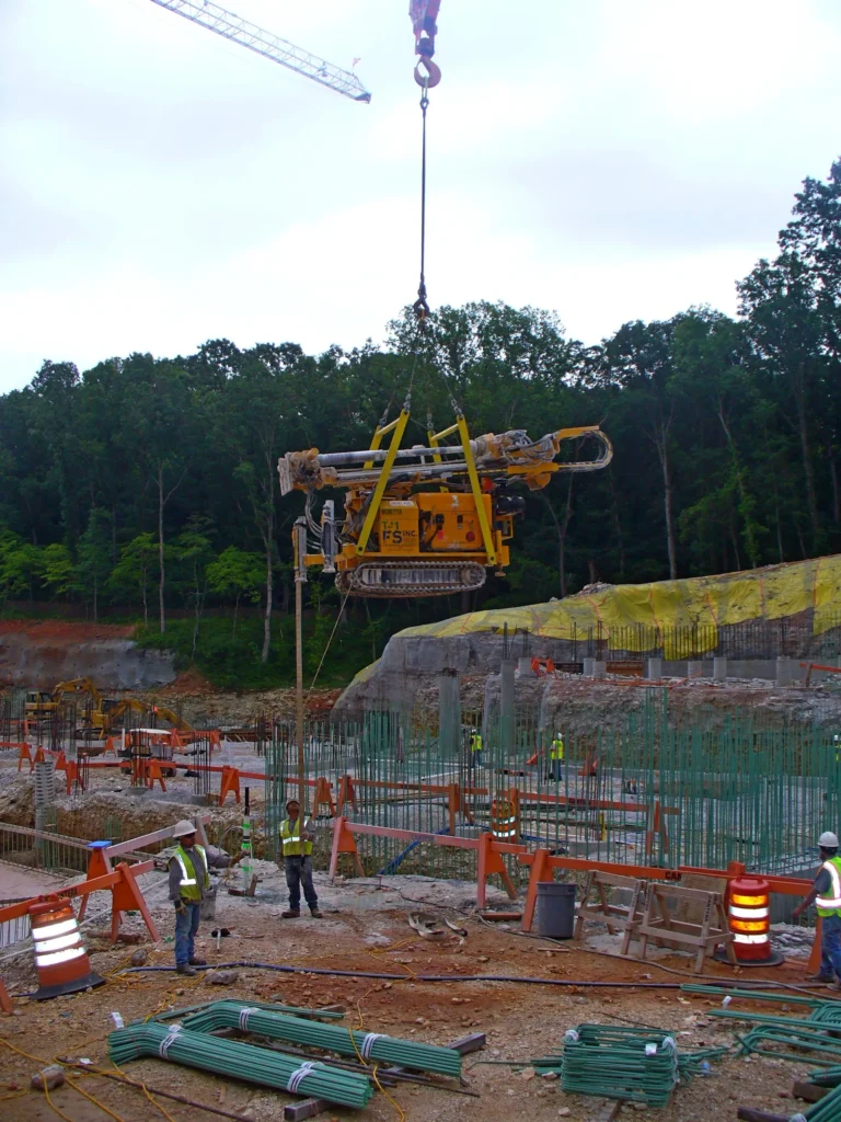 A construction site with a large drill suspended by a crane. Workers in safety gear guide the drill into place. The site has rebar, caution barriers, and materials scattered around. Trees and a cloudy sky form the background.