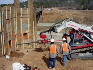 Construction workers operate heavy machinery near a bridge. They are wearing helmets and orange safety vests. A tracked vehicle with a drill attachment is beside them, and wooden structures are set up on the site.