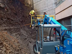 Two construction workers in safety gear are on a blue crane platform drilling into a rocky, vertical surface next to a concrete wall. Debris is scattered below, and one worker steadies a large drill as the other observes.