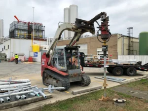 A red skid steer loader is using an auger attachment to drill into the ground at an industrial site. Nearby, workers and various pipes are visible. Industrial buildings and overcast skies form the background.