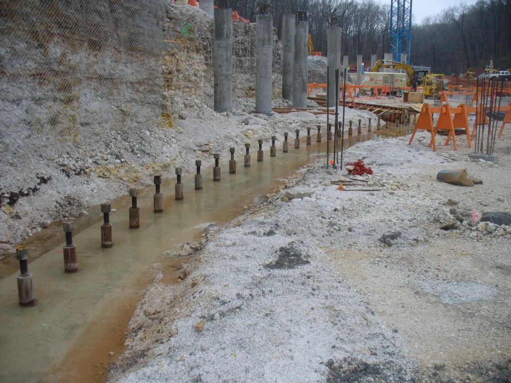 Construction site with rows of concrete pillars along a dug trench. The area is surrounded by dirt and gravel. In the background, there are construction materials, equipment, and a forested area.