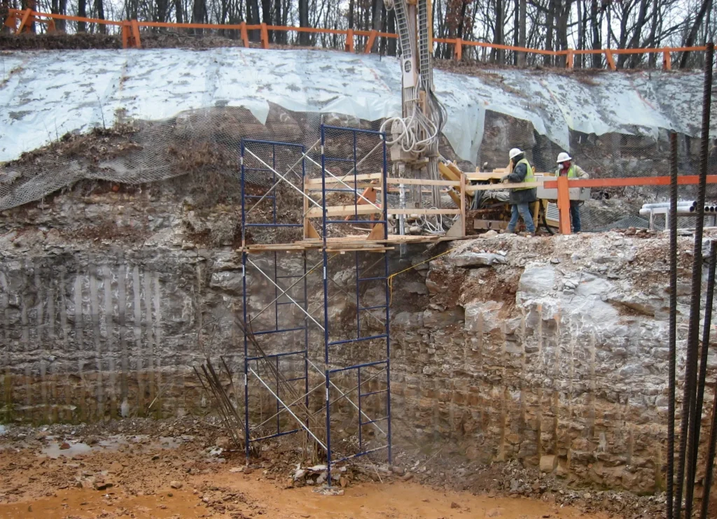 Construction workers operate machinery at a rocky excavation site. They stand on scaffolding near a steep cliff edge, surrounded by trees and soil. Safety barriers are visible at the top of the excavation.