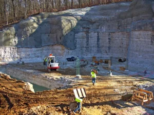 Construction site in a forested area with workers in safety vests. A red excavator is present on the muddy terrain. Workers carry materials, and there are pillars and equipment scattered around. A large rock or soil wall forms the backdrop.