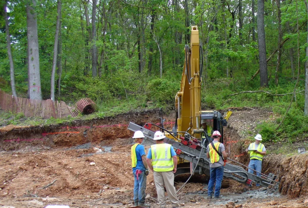 Four construction workers in safety gear stand near heavy machinery at a forested construction site. An excavator is digging in the dirt, surrounded by trees and a barrier fence.