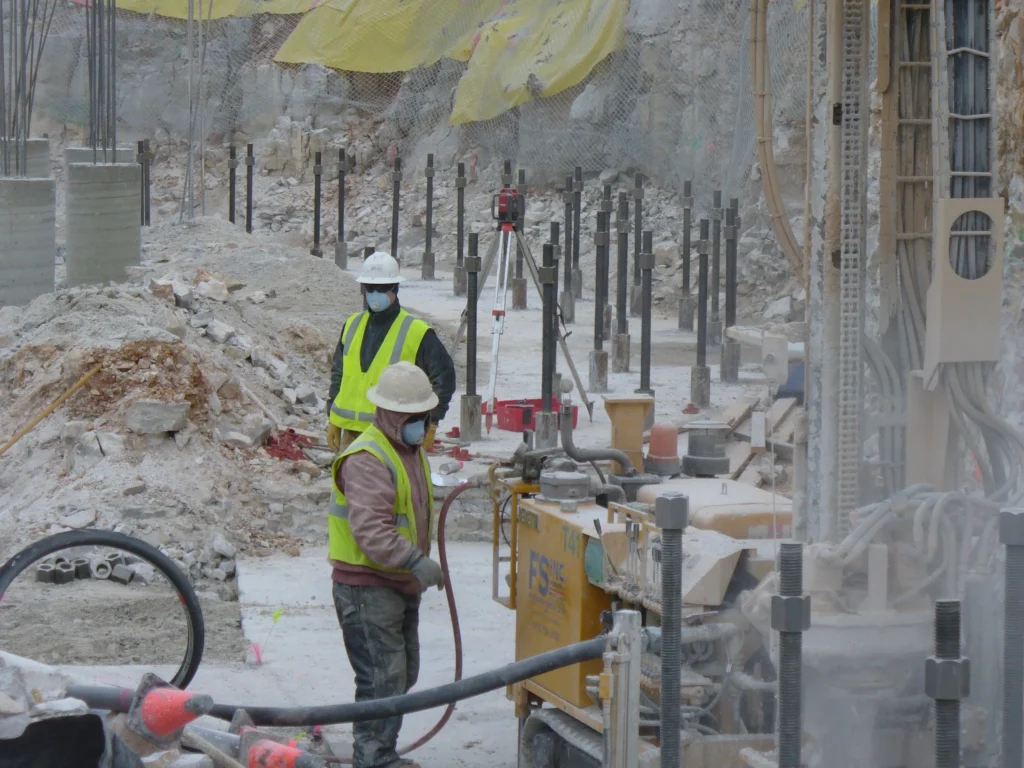 Two construction workers wearing helmets, face masks, and high-visibility vests operate machinery at a construction site. They are surrounded by piles of dirt, metal rods, and a partially erected structure. Dust is visible in the air.