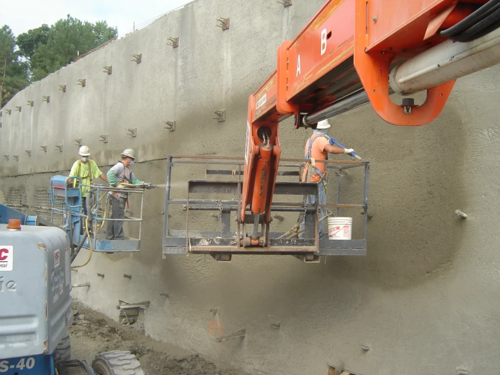 Construction workers wearing safety gear use machinery to apply concrete to a large retaining wall. They are elevated on platforms, and the wall has protruding rebar. Heavy equipment is visible in the foreground, and the ground is muddy.