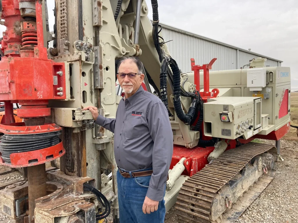 A man wearing glasses and a gray shirt stands beside a large industrial drilling machine. He rests one hand on the machinery, which is red and beige, near a metal building on a gravel area. The sky is overcast.