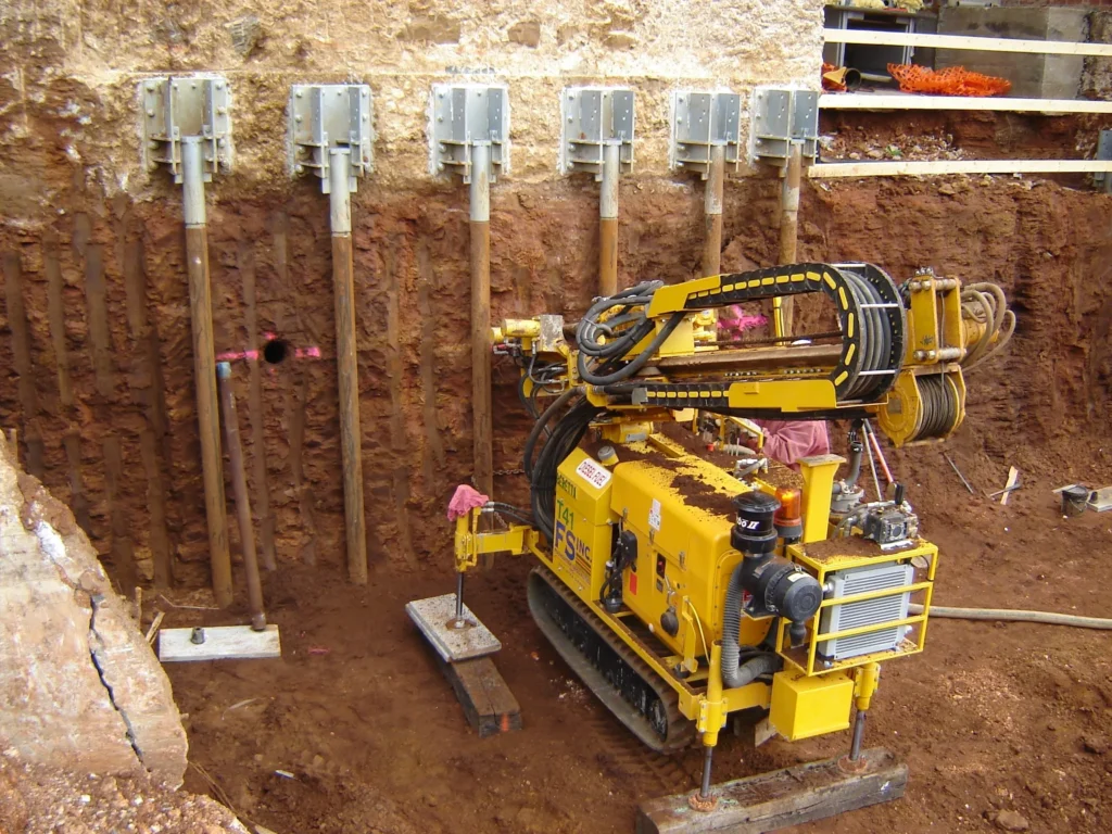 A small yellow drilling machine is in action on a construction site, installing anchors into a dirt and rock wall. Several metal rods are already embedded, creating a stable support structure. Tools and materials are scattered around.