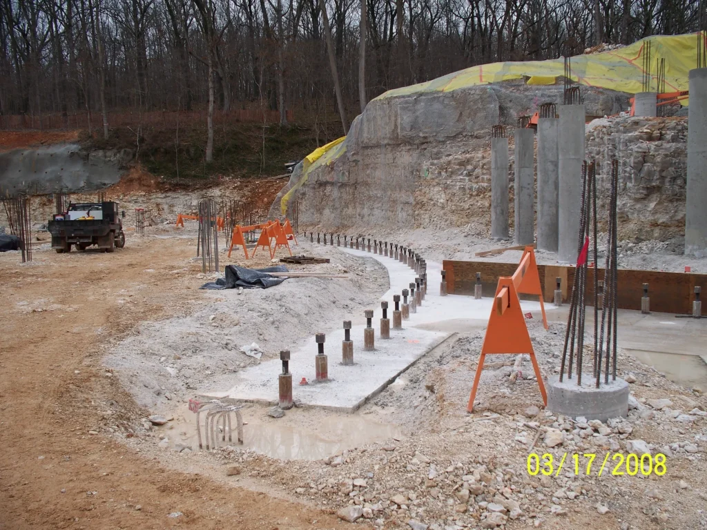 Construction site in a wooded area with concrete pillars, rebar, and orange safety barriers. A pathway lined with metal rods is visible, and equipment is scattered around.