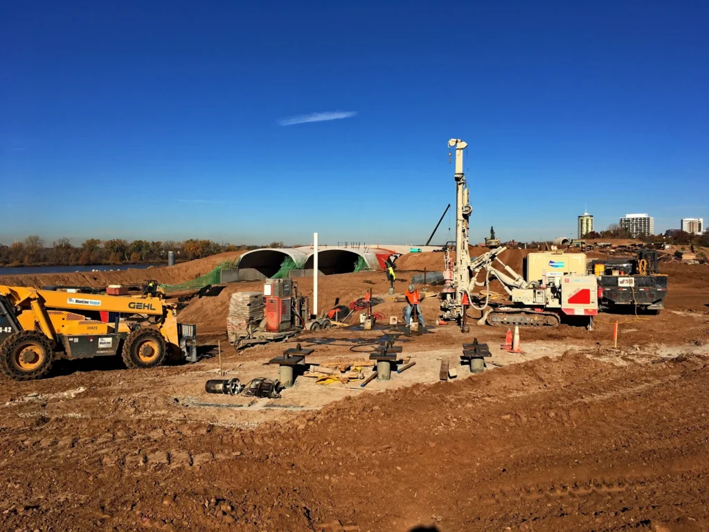 Construction site with machinery and workers on a clear day. Two large vehicles and a pile driver are operating on a dirt lot. Tunnels and city buildings are visible in the background. Orange cones and materials are scattered around.