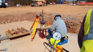 A construction worker in a hoodie and hard hat is operating a hydraulic gauge and other equipment at a construction site. The ground is bare, with machinery and tools scattered around. A tripod and measurement tools are also visible.