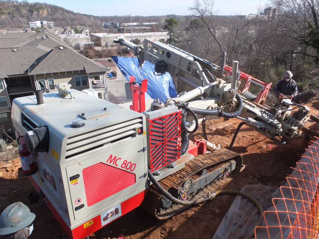 A large drilling machine labeled "MC 800" is positioned on a sloped, rocky construction site. The machine features caterpillar tracks and a complex drilling arm. Nearby are safety fences and residential buildings, with some trees and hills in the background.