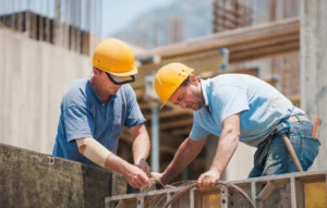 Two construction workers in yellow hard hats focus on wiring at a construction site. They are working together on a wooden framework, surrounded by building materials and scaffolding on a sunny day.