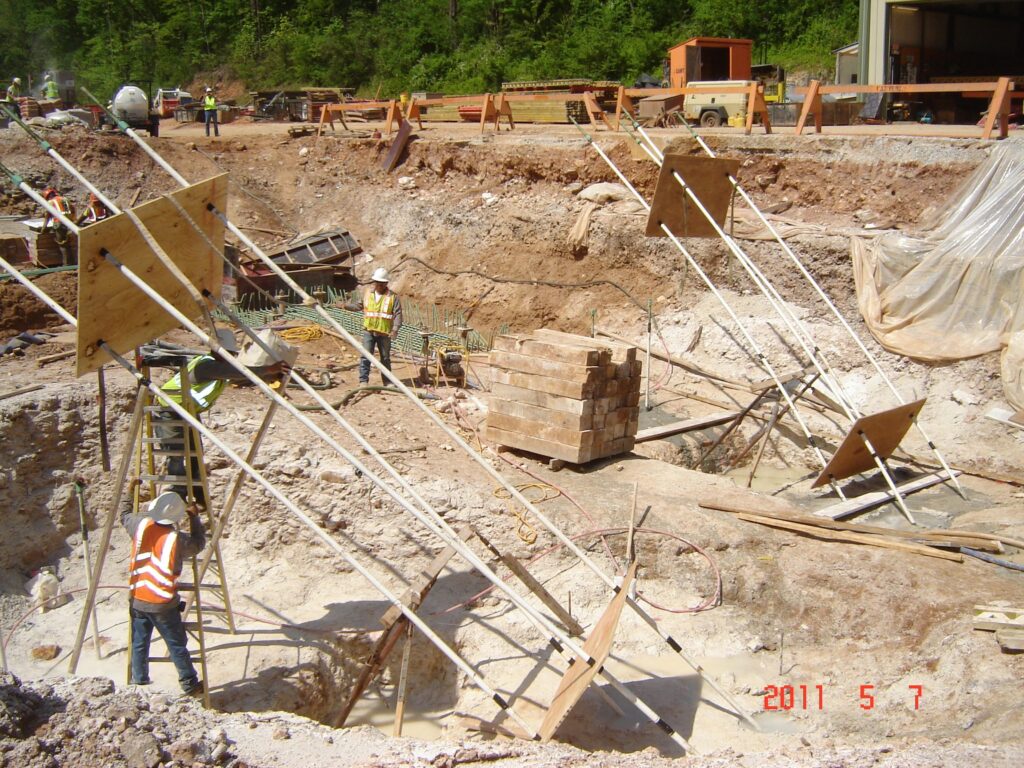 Construction site with workers wearing safety vests and helmets. They are setting up support beams around a deep excavation with a ladder and bricks nearby. Equipment and materials are scattered around the site.