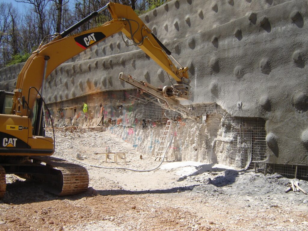 A construction site with a large excavator working on a reinforced concrete wall. The wall has a textured surface with a grid of protruding anchors. A ladder leans against the wall, and construction materials are scattered on the ground.