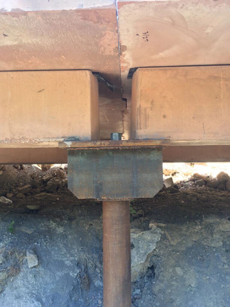 View of a rusted metal support beam under a concrete structure, showing signs of deterioration. The supporting pillar stands on rocky ground, highlighting age and environmental wear.