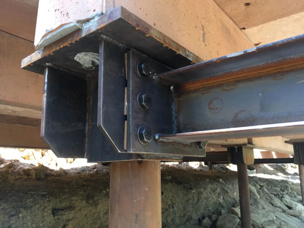 Close-up of a steel beam connection on a construction site. The beam is bolted to a support structure, with visible rust and welding marks. It is positioned atop a vertical wooden support post. The ground below is uneven and earthy.