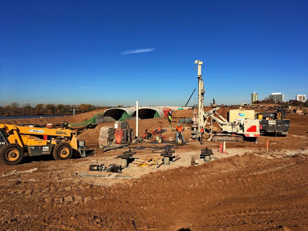 Construction site with heavy machinery, including a Gehl telehandler and a drilling rig, on a dirt landscape. Workers and equipment surround the area under a clear blue sky. Residential buildings are visible in the distance.