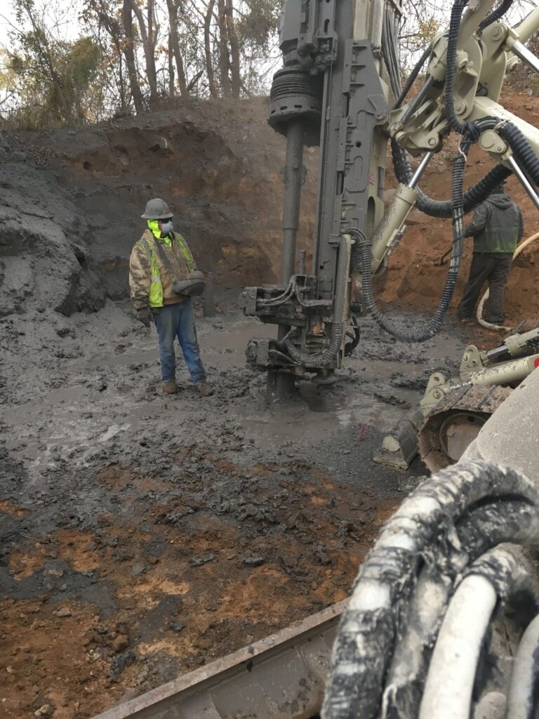 A construction worker stands beside a large drilling machine on a muddy site. The worker is wearing a hard hat, safety glasses, and a high-visibility jacket. The area is surrounded by dirt and trees in the background.