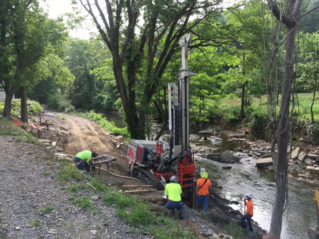 Workers in safety gear operate a large drilling machine near a stream in a forested area. The scene includes dirt paths and lush green trees, indicating a construction project in a natural environment.