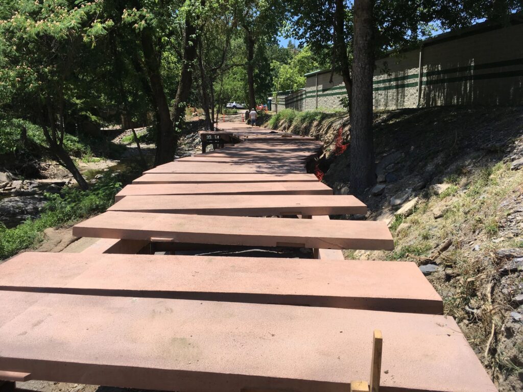 A pathway under construction with large wooden planks spanning across a sloped area beside trees. There is a fence on the right and people in the distance under a clear blue sky.