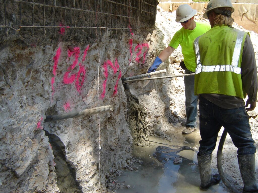 Two construction workers in safety gear inject concrete into a large rock wall with metal reinforcements and red markings. The ground is muddy and wet, and the workers are focused on stabilizing the structure.