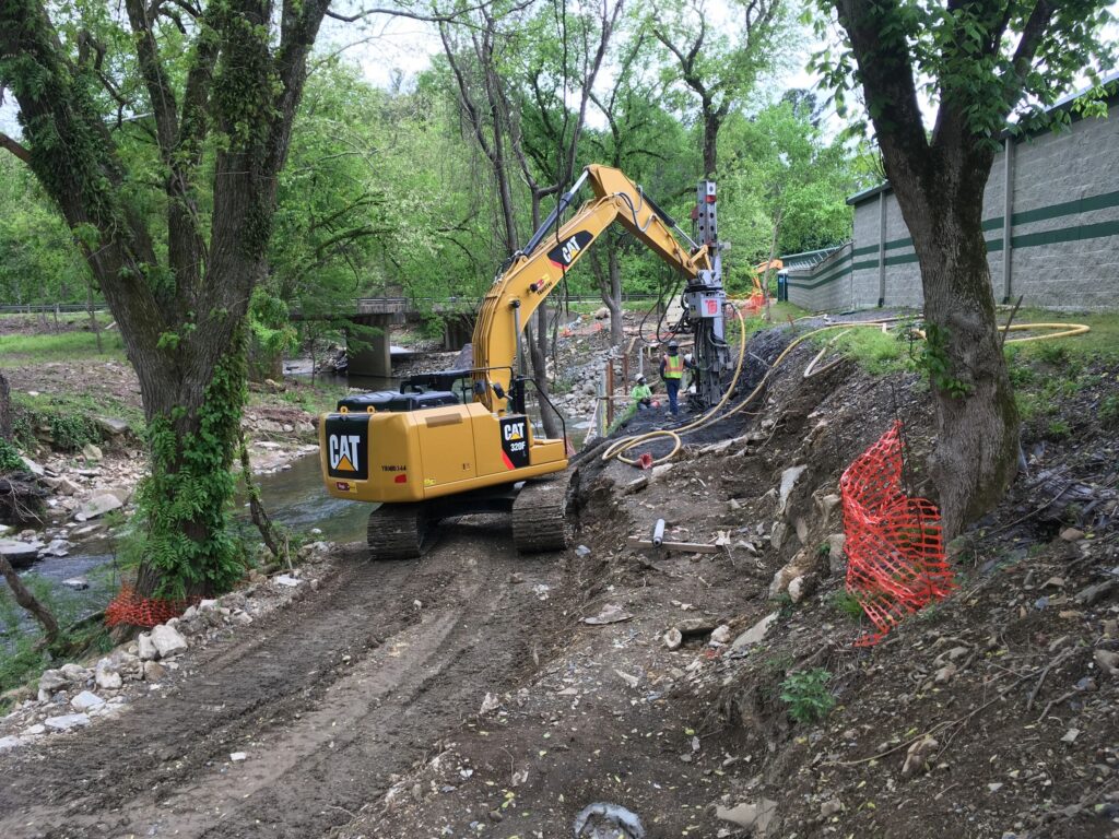 A yellow excavator operates on a dirt path next to a stream surrounded by green trees. Workers are visible near a building, and orange safety netting lines the construction area. Rocks and equipment are scattered around the site.