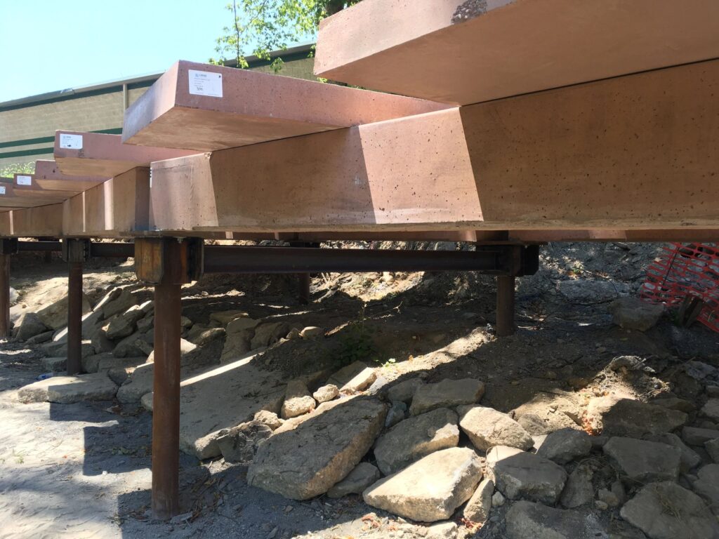 View of large concrete beams supported by steel posts over a rocky ground. The beams are parallel and elevated, creating a shadowed area underneath. A building and foliage are visible in the background.