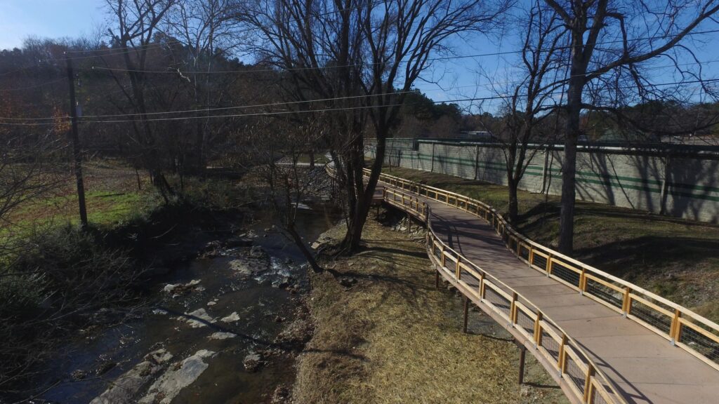 A winding wooden walkway runs alongside a narrow stream in a wooded area. The path is elevated and surrounded by bare trees. A concrete wall is visible on the right, running parallel to the path. The sky is clear and blue.