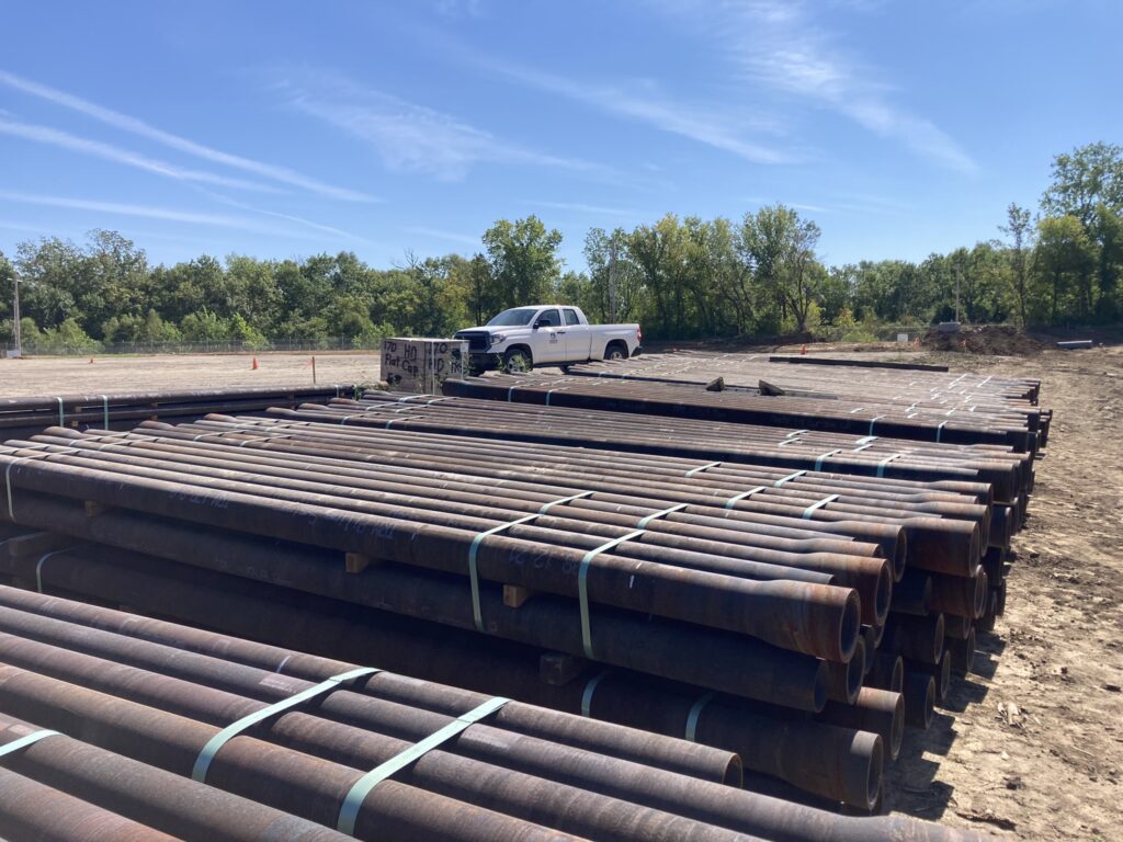 Stacked metal pipes are arranged on a dirt lot with a white pickup truck parked nearby. Trees and a clear blue sky are in the background.