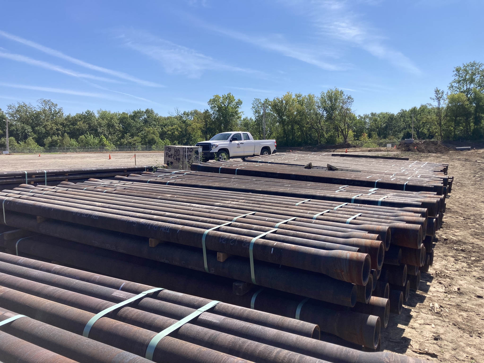 Stacks of large, rusted metal pipes are neatly arranged on the ground at a construction site. A white pickup truck is parked in the background. Trees and a clear blue sky are visible.