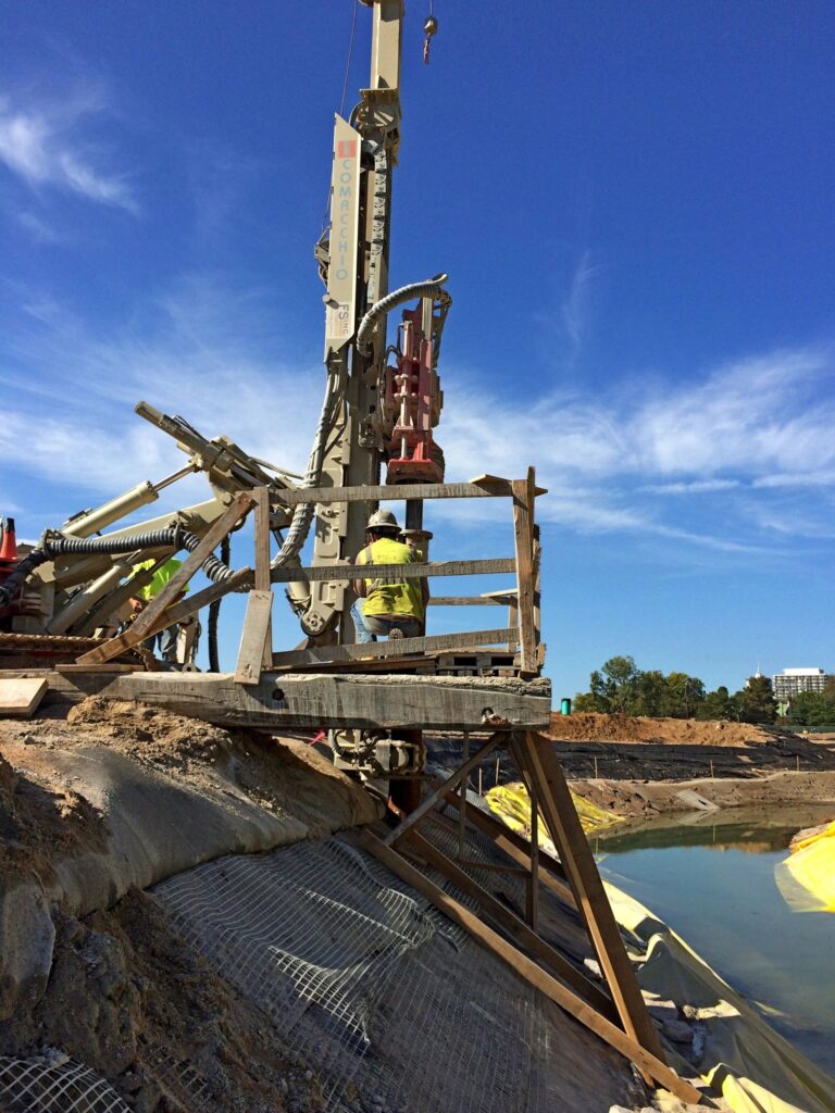 A construction worker operates a large drilling machine on a slope near a water body. The worker is wearing a yellow safety vest and helmet. The sky is clear and blue, and there are trees in the background.