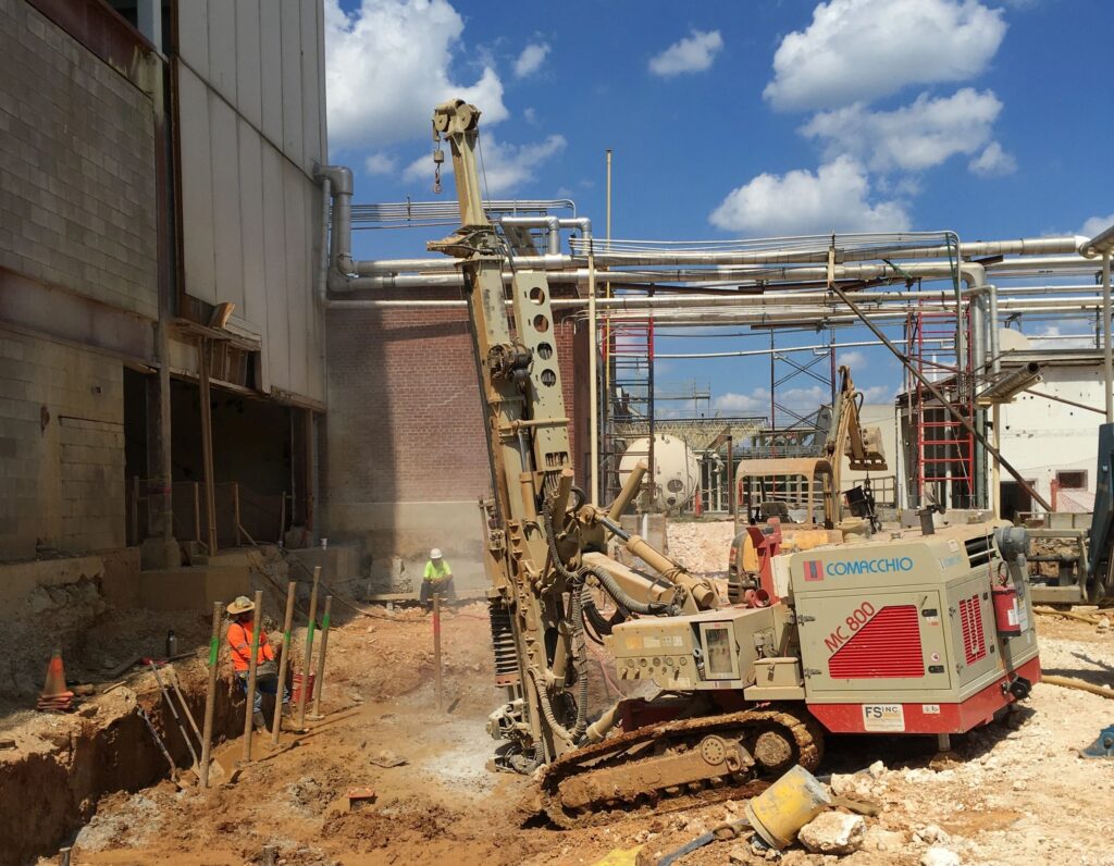 A construction site with a large drilling machine operated by a worker wearing a neon vest and helmet. Nearby, another worker observes. The background shows industrial piping and scaffolding against a blue sky with clouds.