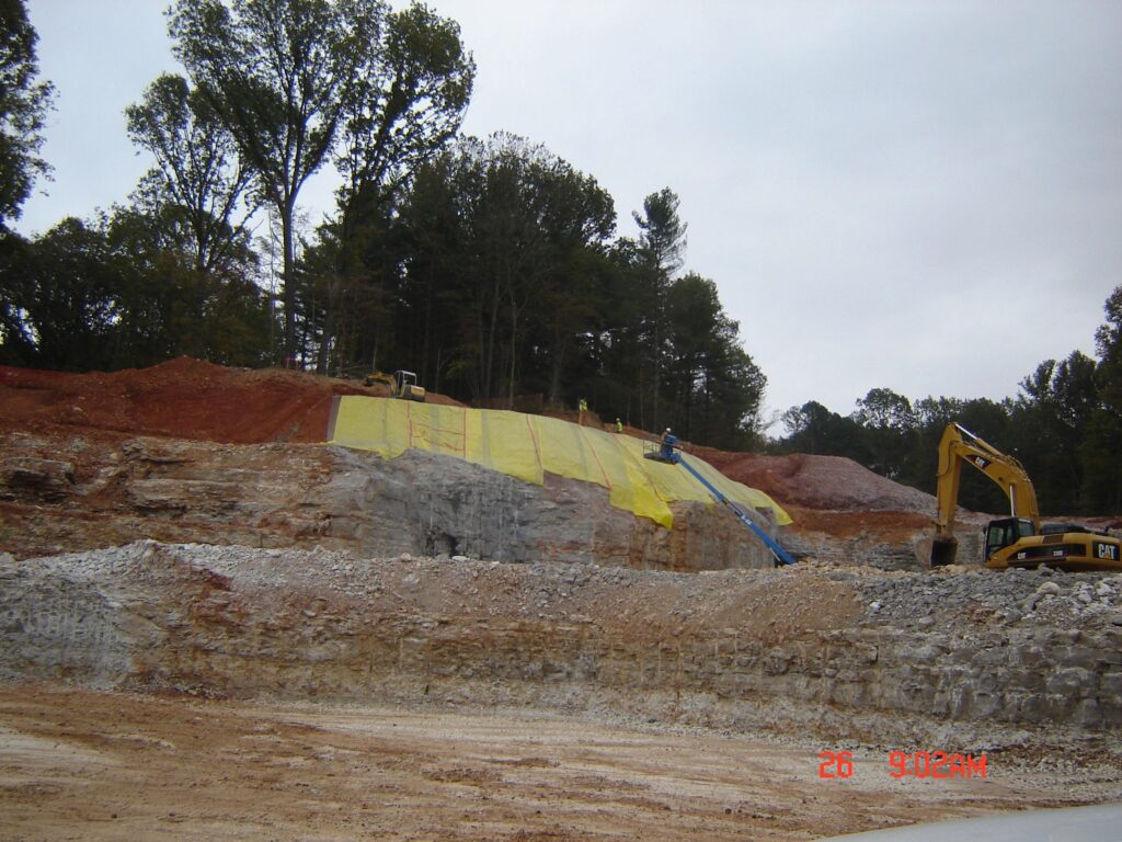 Construction site with workers applying yellow concrete protection on an inclined surface. An excavator operates nearby, surrounded by earth and trees. The scene captures the progress of land preparation and safety measures.