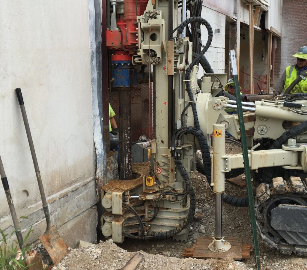 A construction site with drilling equipment positioned next to a building wall. Workers are wearing safety gear. Soil and construction materials are visible, along with a shovel leaning against the wall.