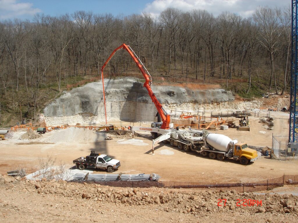 A construction site with a concrete pump truck pouring concrete into a foundation near a cliff. Several vehicles and construction materials are scattered on the ground. Bare trees surround the area under a blue sky with clouds.