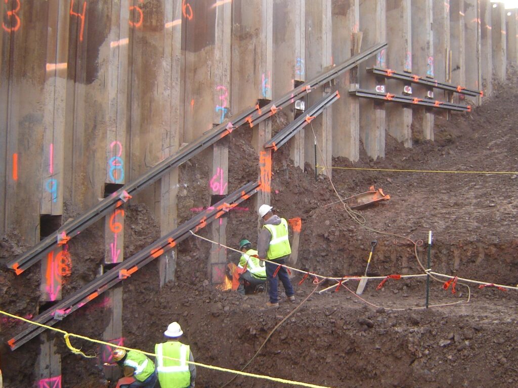 Construction workers wearing safety vests and helmets are working on a large excavation site, reinforcing a concrete retaining wall with metal braces. The wall has colorful numbers and markings, and the ground is muddy.