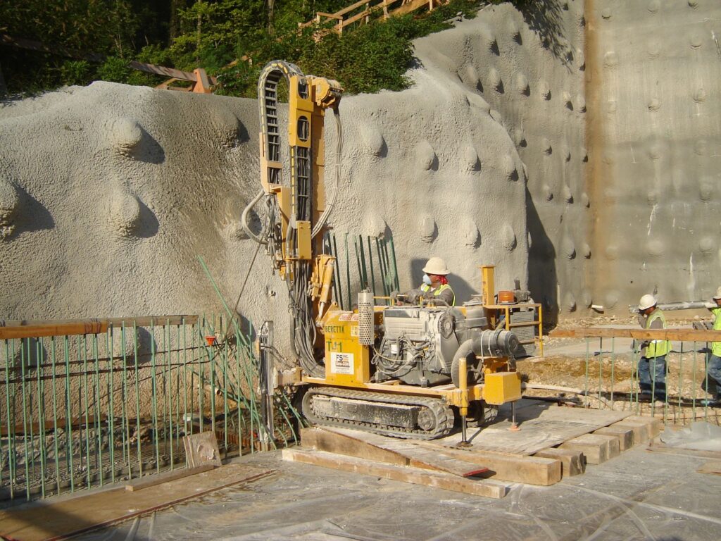 Construction workers operate a large drilling machine near a reinforced concrete wall with visible anchor points. The area is surrounded by safety barriers, with trees in the background, under a sunny sky.