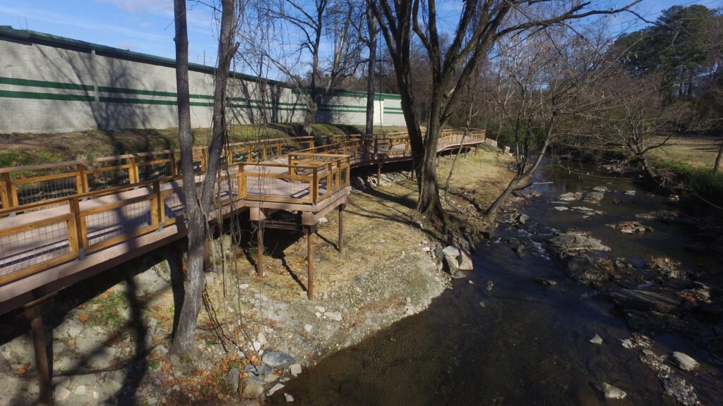 A wooden boardwalk runs alongside a small stream, elevated on stilts above rocky ground. Leafless trees line the path, casting shadows. A concrete wall with green stripes is on the left, and the area appears to be a quiet, natural setting.