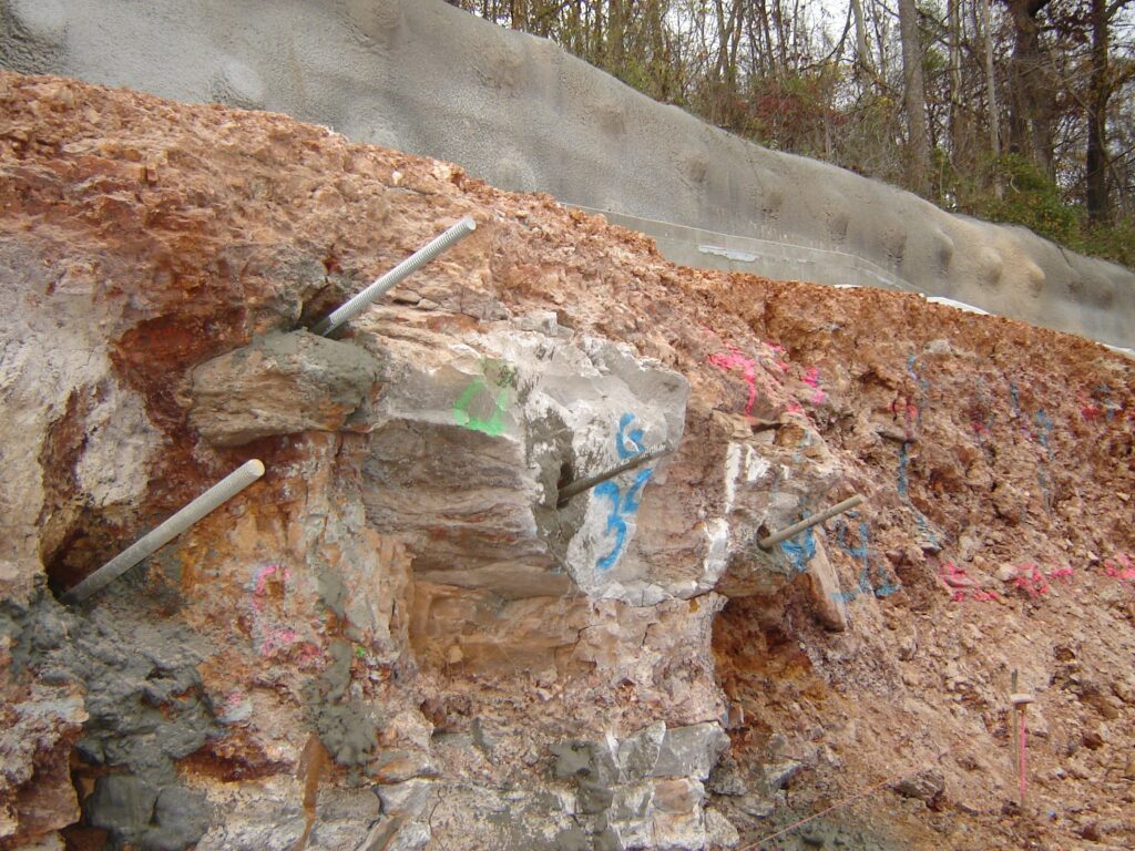 Rock face with exposed soil and visible rebar supports, marked with blue and green spray paint. The area appears to be part of a construction or excavation site, with trees in the background.