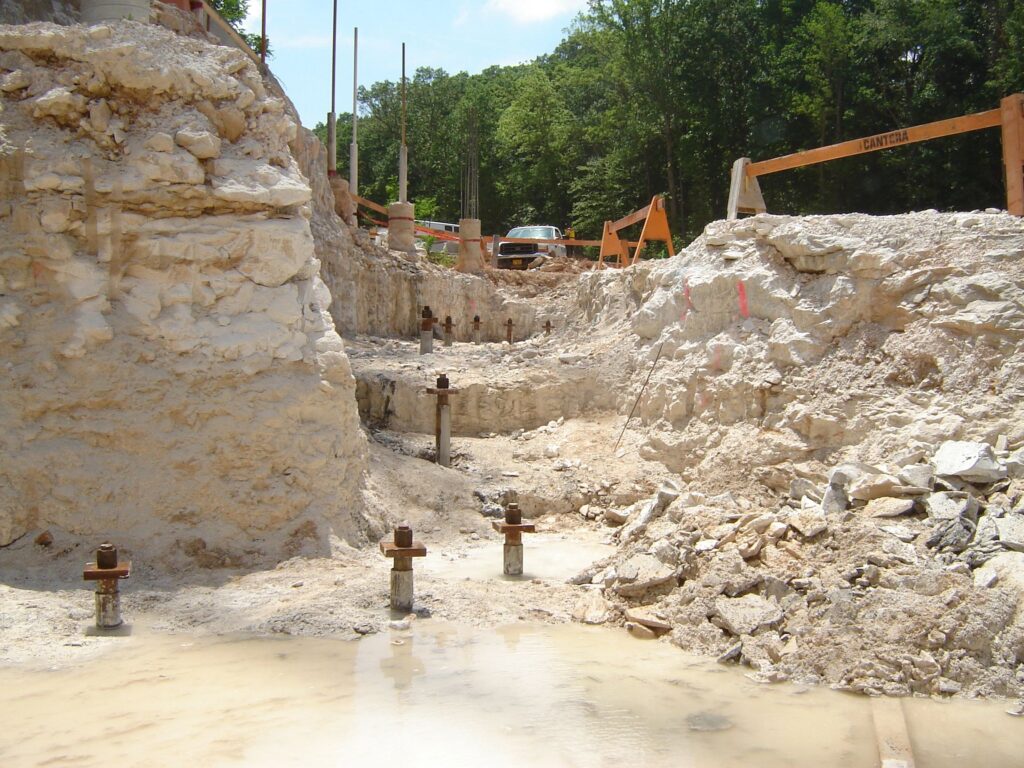 A construction site with rocky terrain and a trench, reinforced with metal rods. There's a muddy puddle in the foreground and an orange vehicle parked above. Trees and caution barriers are visible in the background.