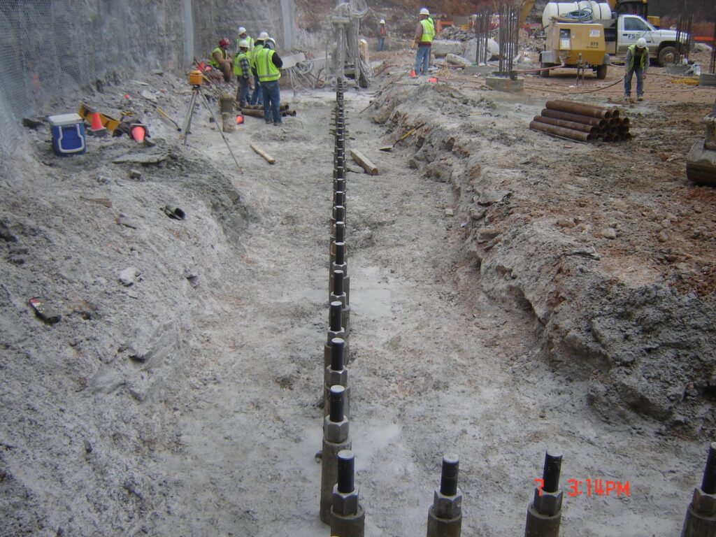 Construction site with several workers in safety gear installing a row of metal rods in a trench. Equipment is scattered around, including pipes and a cement mixer. The ground appears muddy, indicating ongoing foundational work.