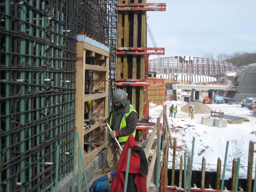 A construction worker in safety gear stands on a snowy site, using a tool on a wooden and metal structure. In the background, other workers and construction frameworks are visible under a partly cloudy sky.