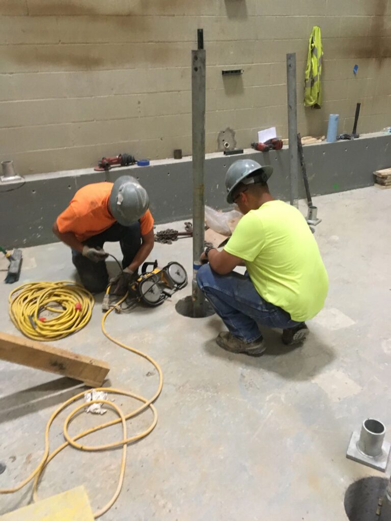 Two construction workers in hard hats work on metal poles in a construction site. One is using a power tool with coiled cables nearby. Safety gear and building materials are visible in the background.