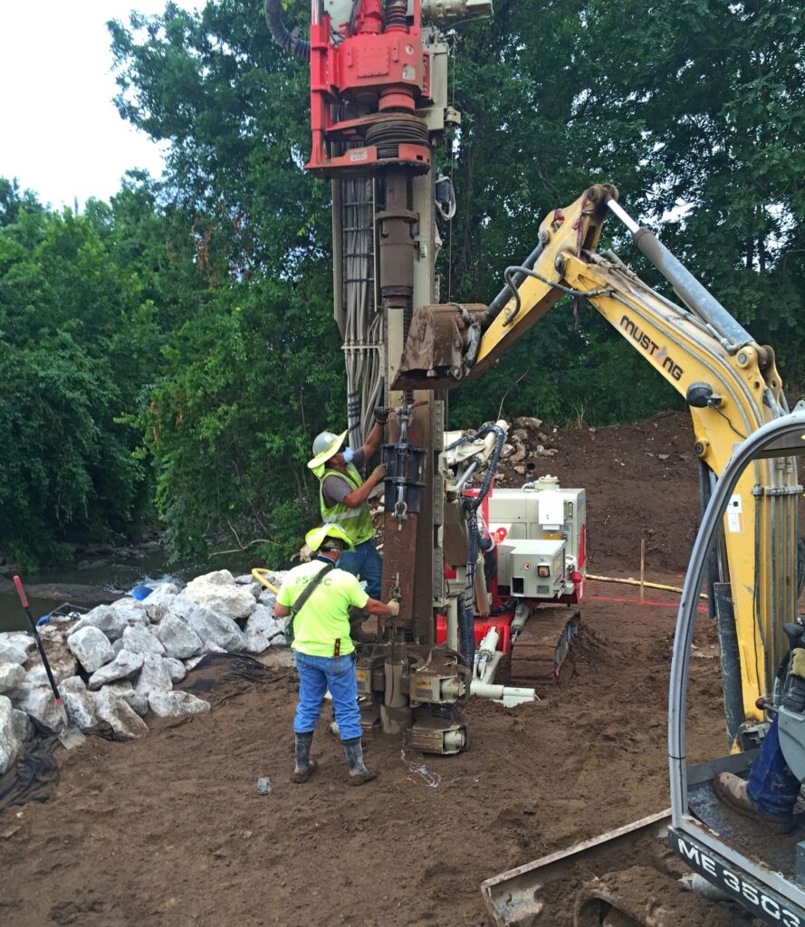 Workers operate heavy machinery for drilling at a construction site surrounded by dirt and rocks. They're wearing safety gear, including helmets and reflective vests. Trees line the background.